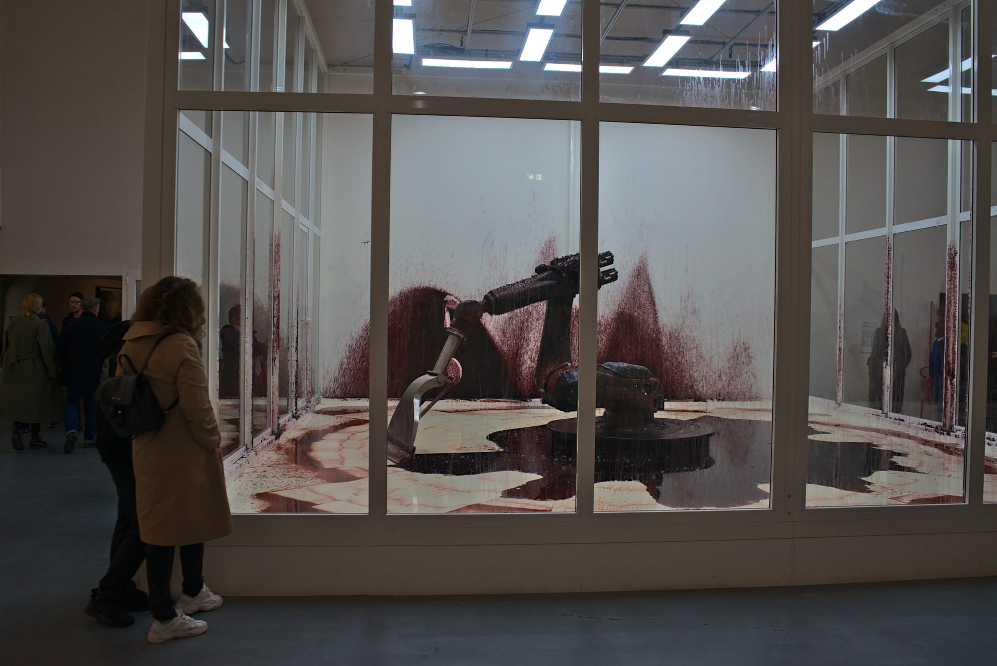 A robot is inside a glass chamber with people looking on the outside. The robot is covered in blood, a puddle forming next to it. The robot scoops the blood towards itself, occasionally getting it on the walls of the chamber.
